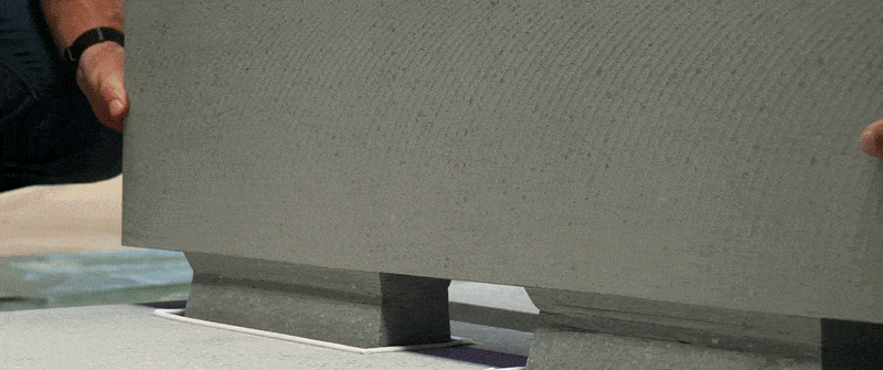 Ein Mitarbeiter in Schutzkleidung und Handschuhen füllt zur Konsistenzprüfung im Labor einen zähflüssigen Carbonbeton in ein Prüfgefäß. Der Beton ist grau und hat eine zähflüssige Konsistenz, während der Mitarbeiter den Eimer vorsichtig über dem Prüfgefäß hält, um den Beton hineinzufüllen. Die Szene spielt in einem Labor oder einer Produktionsumgebung, mit unscharfem Hintergrund, der die Aufmerksamkeit auf die Aktion des Mitarbeiters und das Prüfgefäß lenkt.